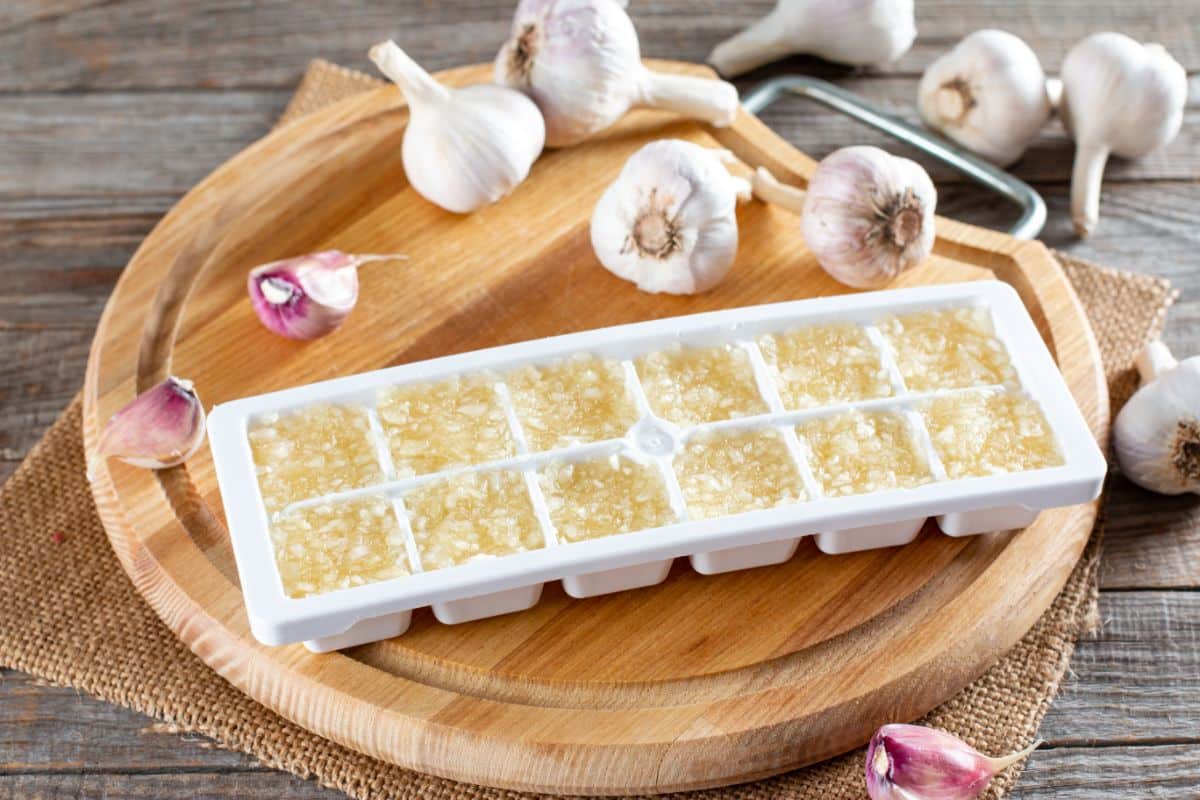 Frozen garlic in a white try on a wooden cutting board with garlic heads scattered around.