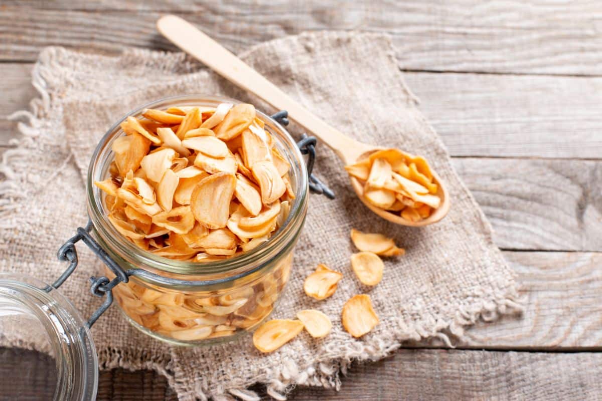 A glass jar with sliced dried garlic on a wooden table with a wooden spoon.